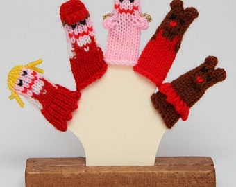 May include: Five knitted finger puppets on a wooden stand. The puppets include a girl with blonde hair, a girl with red hair, a girl with pink hair, and two bears. The puppets are all wearing red and white clothing.