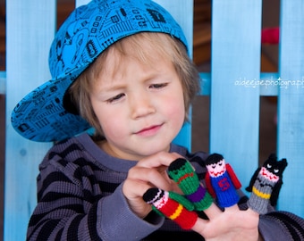 May include: A young person wearing a blue and white patterned hat is playing with finger puppets. The puppets are a green superhero, a red superhero, a blue superhero, and a gray and black batman.