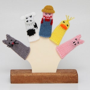 May include: Five finger puppets on a wooden stand. The puppets are a cow, a farmer, a duck, a pig, and a cat. The puppets are all knitted and have different colors and patterns.