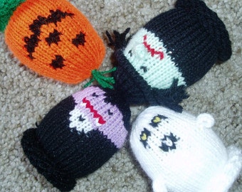 May include: Four knitted Halloween characters: a pumpkin, a vampire, a ghost, and a witch. The pumpkin is orange with a black jack-o-lantern face. The vampire is purple and black with a red mouth. The ghost is white with black eyes. The witch is green and black with a red nose.