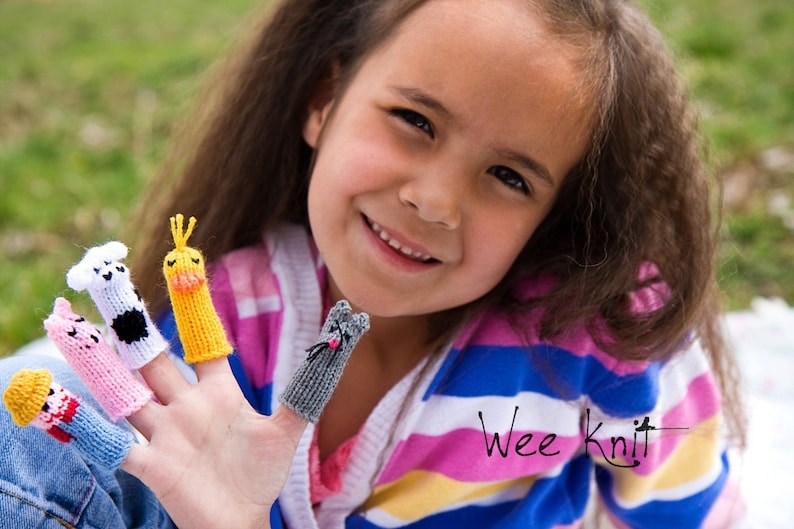 May include: A young girl wearing a pink and blue striped shirt with the text "Wee Knit" is wearing finger puppets on her fingers. The finger puppets are a cow, a chick, a pig, and a cat.