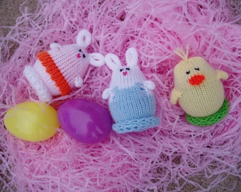 May include: Three knitted Easter decorations: a white bunny with an orange stripe, a blue bunny with a white stripe, and a yellow chick with a green stripe. The decorations are sitting on a bed of pink shredded paper.