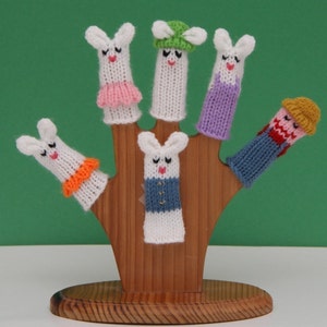 May include: Five finger puppets on a wooden stand. The puppets are all white with different colored accents. One puppet has a green hat, one has a purple dress, one has an orange collar, one has a blue and white shirt, and one has a blue and red outfit.
