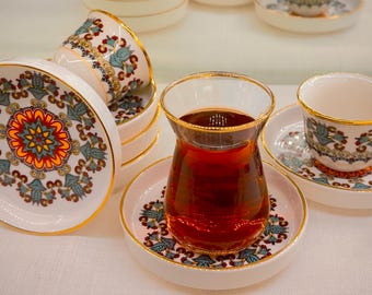 May include: A set of Turkish teacups and saucers with intricate floral patterns in shades of blue, red, and gold. The cups are made of white porcelain and have a gold rim. The saucers have a matching design and are also made of white porcelain. A glass of dark tea sits on a saucer.