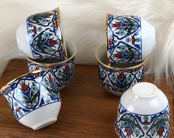 May include: A set of six small, white ceramic cups with a vibrant floral design in blue, red, and green. Each cup has a gold rim and is arranged on a wooden tray, ideal for serving coffee or espresso.