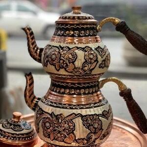 Könnte beinhalten: Eine zweistöckige Kupferteekanne mit aufwendigen floralen Mustern und schwarzen Akzenten. Die Teekanne hat einen Holzgriff und einen Deckel mit ähnlichem Design. Die Teekanne steht auf einem Kupfertablett.