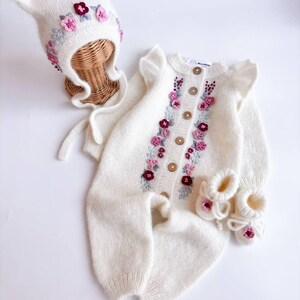 May include: A white knitted baby outfit set, including a hat, romper, and booties. The set is adorned with embroidered pink and burgundy flowers. The romper has wooden buttons and ruffled sleeves. The hat has cat-like ears.