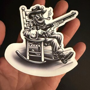 Puede incluir: Adhesivo en blanco y negro que representa a un hombre barbudo con sombrero de vaquero, fumando en pipa y sosteniendo una escopeta, sentado en una mecedora. Una botella marcada con "XXX" est&aacute; a su lado. Borde blanco.