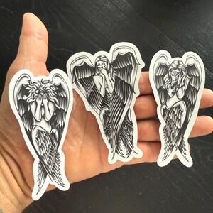 May include: Three black and white angel stickers with detailed wing designs. Each sticker depicts an angel in a different pose, with one covering their face, another covering their mouth, and the last looking down.