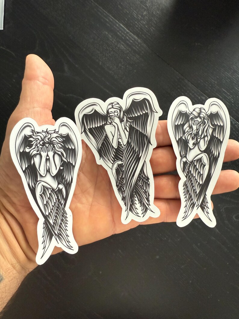 May include: Three black and white angel stickers with detailed wing designs. Each sticker depicts an angel in a different pose, with one covering their face, another covering their mouth, and the last looking down.