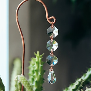 May include: A copper wire plant stake with a crystal prism hanging from it. The prism is made up of five clear hexagonal crystals and a teardrop-shaped crystal.