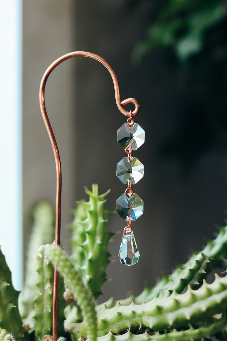 May include: A copper wire plant stake with a crystal prism hanging from it. The prism is made up of five clear hexagonal crystals and a teardrop-shaped crystal.