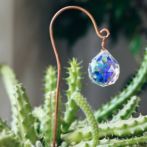 May include: A copper wire plant stake with a crystal prism hanging from it. The stake is placed in a pot of green cactus plants.