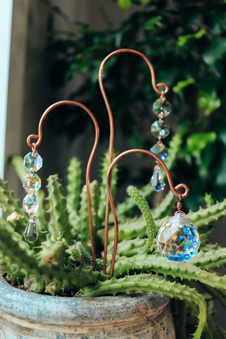 May include: Three copper wire garden stakes with crystal prisms hanging from them. The stakes are inserted into a pot of green succulents.