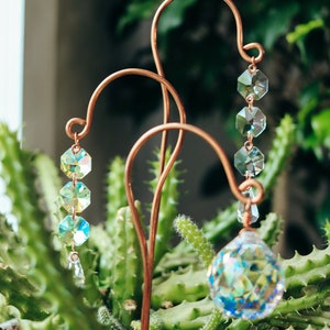 May include: A copper wire garden stake with three crystal prisms hanging from it. The stake is placed in a pot of green cactus plants.