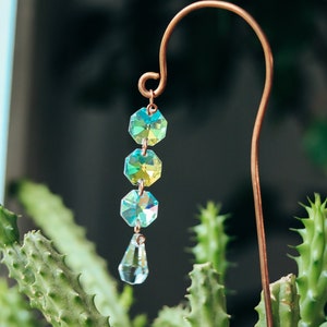 May include: A copper plant stake with a crystal prism hanging from it. The prism is made of clear glass with iridescent green and blue hues. The stake is inserted into a green cactus plant.