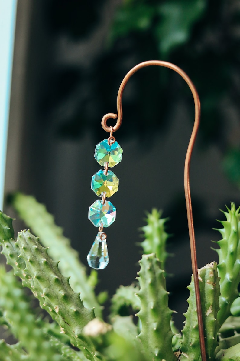 May include: A copper plant stake with a crystal prism hanging from it. The prism is made of clear glass with iridescent green and blue hues. The stake is inserted into a green cactus plant.