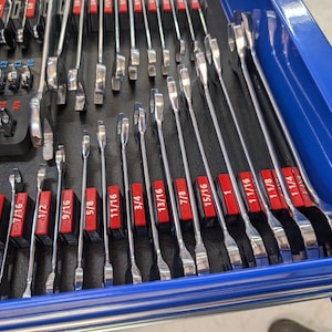 May include: A collection of shiny, silver wrenches of various sizes, neatly organized in a blue toolbox drawer. Each wrench is labeled with its size in inches, displayed on red blocks. The tools are arranged for easy access.