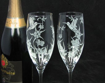 May include: Two clear glass champagne flutes with etched hummingbird designs and floral patterns. A bottle of champagne with a gold label is in the background. A pearl necklace and two gold rings are in the foreground.