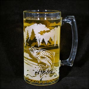 May include: A clear glass beer mug with a handle, etched with a trout jumping out of the water. The mug is filled with a light yellow beer and has a forest scene etched on the side.