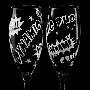 Two clear glass champagne flutes with etched comic book style designs. One flute says "Dynamic Duo" and the other says "CRASH WHAM!" with a "POW!" graphic.