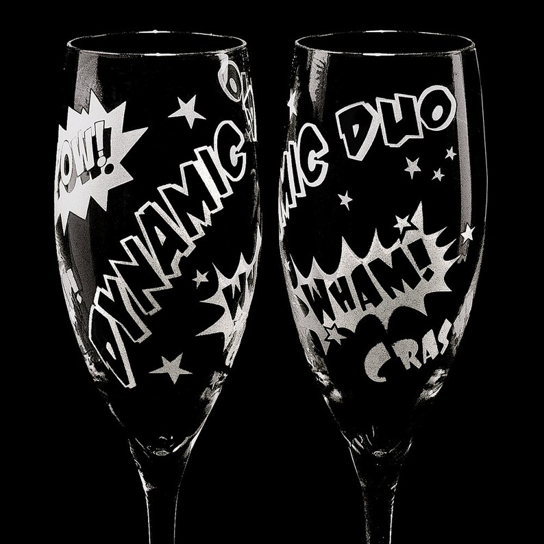 Two clear glass champagne flutes with etched comic book style designs. One flute says "Dynamic Duo" and the other says "CRASH WHAM!" with a "POW!" graphic.