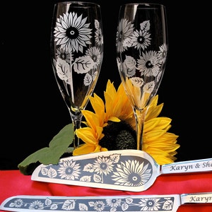 May include: Two champagne flutes with etched sunflower designs and a matching cake knife with the text "Karyn & Shane" and "Karyn" on the handles. The flutes and knife are on a red surface with a yellow sunflower in the background.