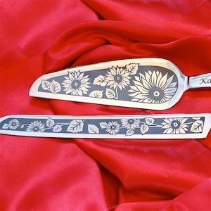 May include: A set of two silver cake knives with sunflower designs. The knives have a black handle and the words "Ka" and "Sa" are engraved on the handles.