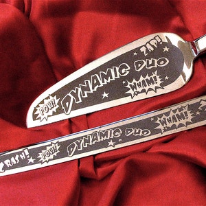 A stainless steel cake server and knife set with etched designs in comic book style. The flutes are engraved with the words "Dynamic Duo" and "Pow! Wham! Crash!"