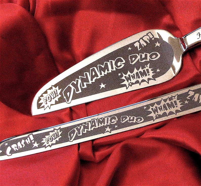 A stainless steel cake server and knife set with etched designs in comic book style. The flutes are engraved with the words "Dynamic Duo" and "Pow! Wham! Crash!"