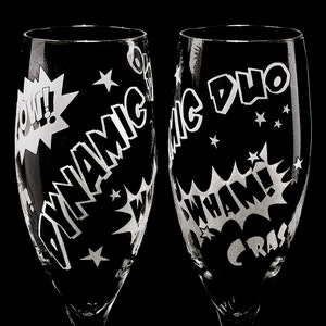 Two clear glass champagne flutes with etched comic book style designs. One flute says "Dynamic Duo" and the other says "CRASH WHAM!" with a "POW!" graphic.