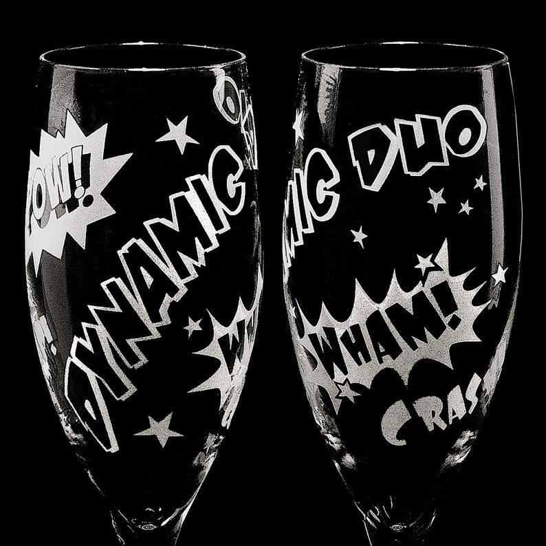 Two clear glass champagne flutes with etched comic book style designs. One flute says "Dynamic Duo" and the other says "CRASH WHAM!" with a "POW!" graphic.