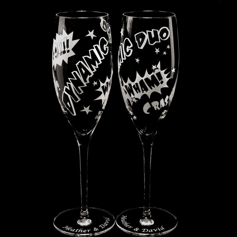 Two clear glass champagne flutes with etched comic book style designs. One flute says "Dynamic Duo" and the other says "CRASH WHAM!" with a "POW!" graphic.  names and wedding date engraved on the flute bases.