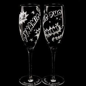 Two clear glass champagne flutes with etched designs in comic book style. The flutes are engraved with the words "Dynamic Duo" and "Pow! Wham! Crash!"