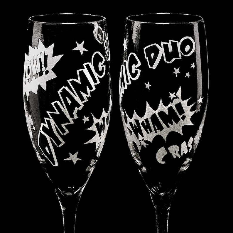 Two clear glass champagne flutes with etched designs in comic book style. The flutes are engraved with the words "Dynamic Duo" and "Pow! Wham! Crash!"