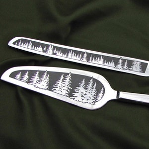 May include: A set of two silver cake knives with a forest scene etched into the blades. The knives are on a dark green fabric background.