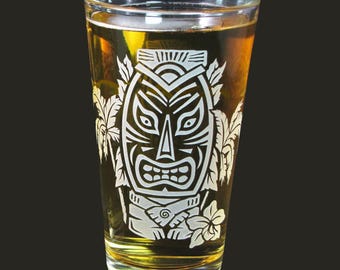 May include: A clear glass pint glass with a white etched design of a tiki mask with palm trees and a flower. The glass is filled with a golden yellow liquid.