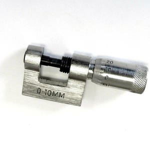 Puede incluir: Un micrómetro plateado con un agarre moleteado y un tornillo negro. El dispositivo está sobre una base rectangular con el texto "0-10MM" grabado. El micrómetro mide de 0 a 20 mm.