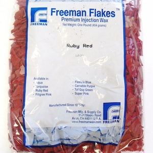 May include: A bag of red Freeman Flakes Premium Injection Wax, labeled "Ruby Red" and weighing one pound (454 grams). The bag is made of clear plastic and shows the wax flakes inside. The label also lists other available colors: Aqua, Turquoise, Filigree Pink, Flexible Blue, Carvable Purple, Tuf Guy Green, and Super Pink. The label states "Manufactured Since 1975 by: Freeman Mfg. & Supply Co. 1101 Moore Road Avon, OH 44011 U.S.A. www.freemanwax.com"
