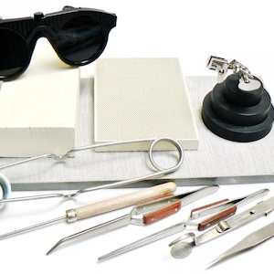 Op de afbeelding: Een set gereedschap voor het maken van sieraden, waaronder een veiligheidsbril, een keramische tegel, een soldeerboutstandaard, een pincet, een tang, een soldeerboutpunt en een klein metalen bekertje.