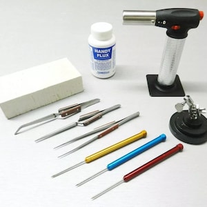 Peut inclure: Un ensemble d'outils de fabrication de bijoux comprenant un chalumeau, un bloc de brique réfractaire, des pinces et une station de soudage. Les outils sont disposés sur une surface blanche. Une bouteille de "Handy Flux" est également incluse.