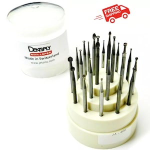 May include: A selection of dental burs in a cream-coloured holder, with a clear plastic container behind. The burs are various shapes and sizes, made of silver metal. The container has the text "DENTSPLY MAILLEFER Made in Switzerland".