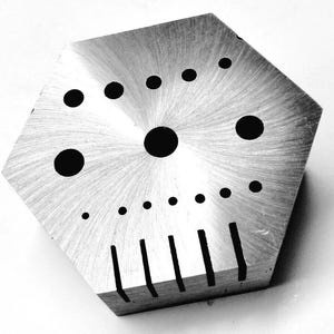 Puede incluir: Una herramienta hexagonal de plata con una variedad de agujeros circulares y ranuras. La herramienta tiene un acabado cepillado y está diseñada para la elaboración artesanal y la fabricación de joyas.