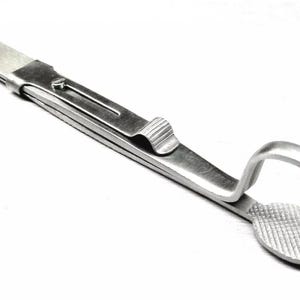 Op de afbeelding: Roestvrijstalen tandheelkundig instrument met een getextureerd, ovaalvormig uiteinde en een gebogen haak. De tool heeft een scharnierend ontwerp met een vergrendelingsmechanisme en een platte, rechthoekige handgreep.