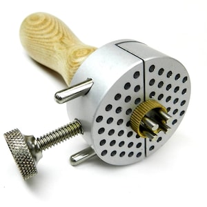 May include: A silver-colored jewelry tool with a wooden handle. The tool has a round, perforated head with a brass-colored ring and several metal posts. A threaded screw with a textured knob is visible.