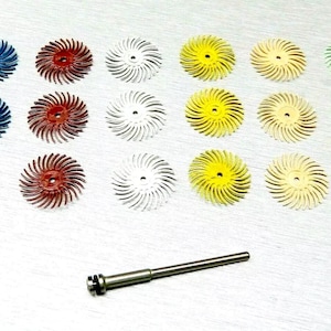 Puede incluir: Un juego de 18 ruedas de lijado en varios colores, incluidos azul, rojo, blanco, amarillo y verde. Las ruedas están dispuestas en un patrón de cuadrícula sobre una superficie plateada. También se incluye un eje de metal.