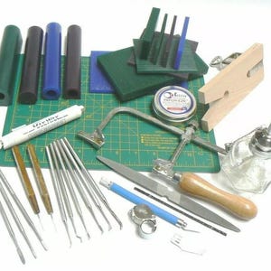 Op de afbeelding: Een set gereedschap voor het maken van sieraden, waaronder een zaag, vijlen, een ringmandrel, een set wax-carving tools, een groene snijmat en een fles wax.