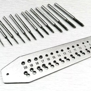 May include: A set of silver metal taps and a tap drill gauge. The taps are cylindrical with pointed ends. The gauge is rectangular with numbered holes of varying sizes, ranging from 1 to 20.