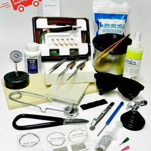May include: A soldering kit containing a range of tools, such as a soldering iron, flux, solder, tweezers, and safety goggles. The kit also features a small crucible, a pair of tongs, and a small bottle of flux. The kit is displayed on a white surface.