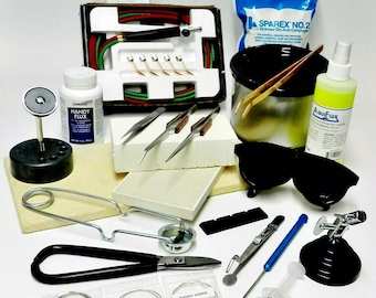 Può includere: Un kit di saldatura con una varietà di strumenti, tra cui un saldatore, flusso, saldatura, pinzette e occhiali di sicurezza. Il kit include anche un piccolo crogiolo, un paio di pinze e una piccola bottiglia di flusso. Il kit è disposto su una superficie bianca.